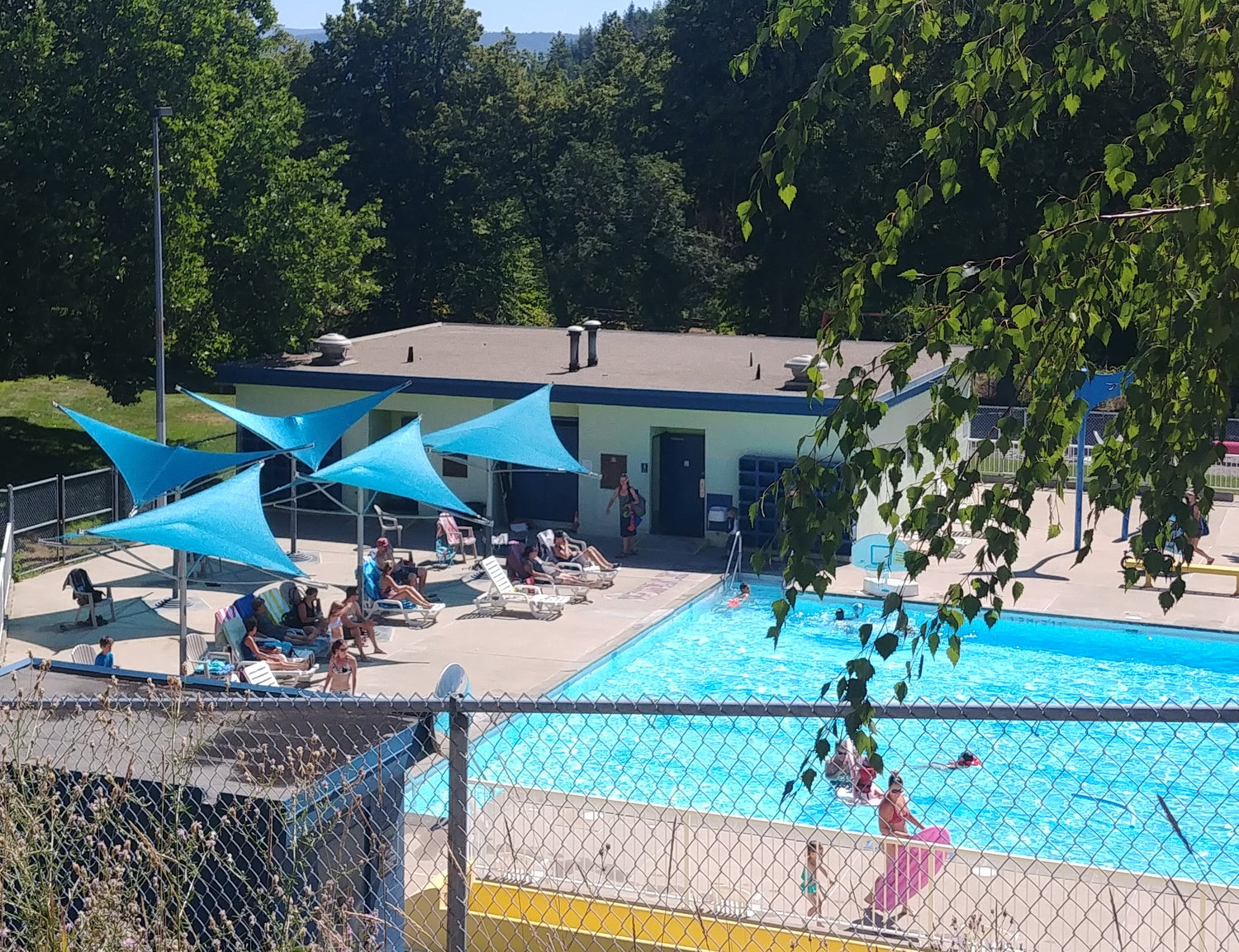 An outdoor swimming pool area featuring several sun shade posts with blue sails providing shade for people lounging on chairs. The sun shade posts create a cool, comfortable environment by the pool, illustrating their effectiveness in outdoor spaces.