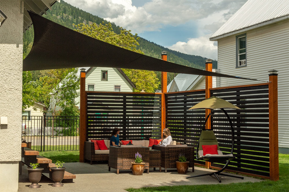 Two people relax under a sleek, black triangle shade sail in a modern backyard setup, showcasing the functionality and style of Standard Sized Shade Sails.