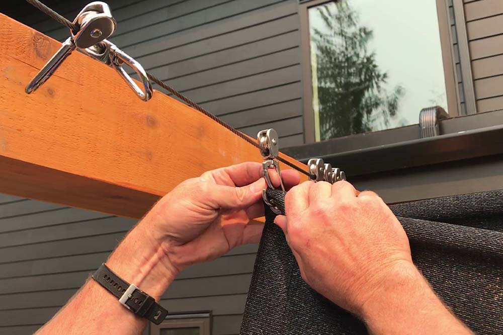 Outdoor Shades installation close-up showing hands attaching a shade cloth using metal clips and tension hardware on a patio frame.