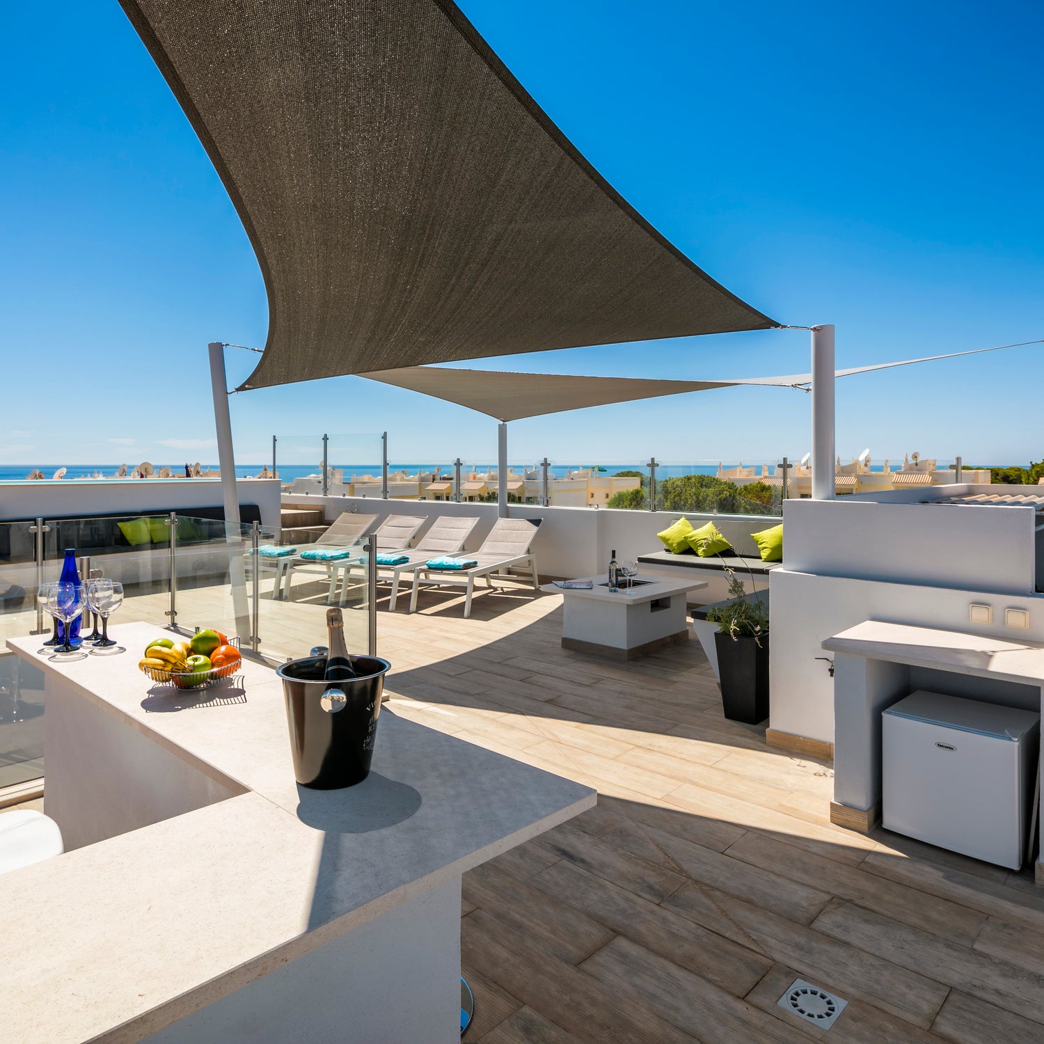Image of a modern rooftop deck with a bar and deck chairs. There are two sundeck covers which are white and dark grey shading the area.