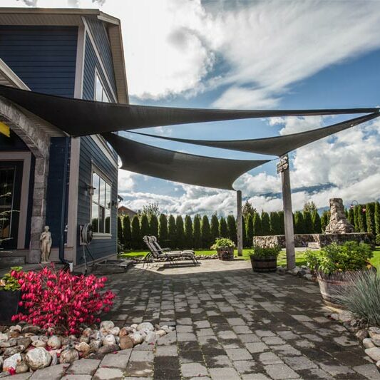 A backyard of a modern house with a stone patio and greenery surrounding it. The image showcases dark grey backyard shade sails covering the patio.