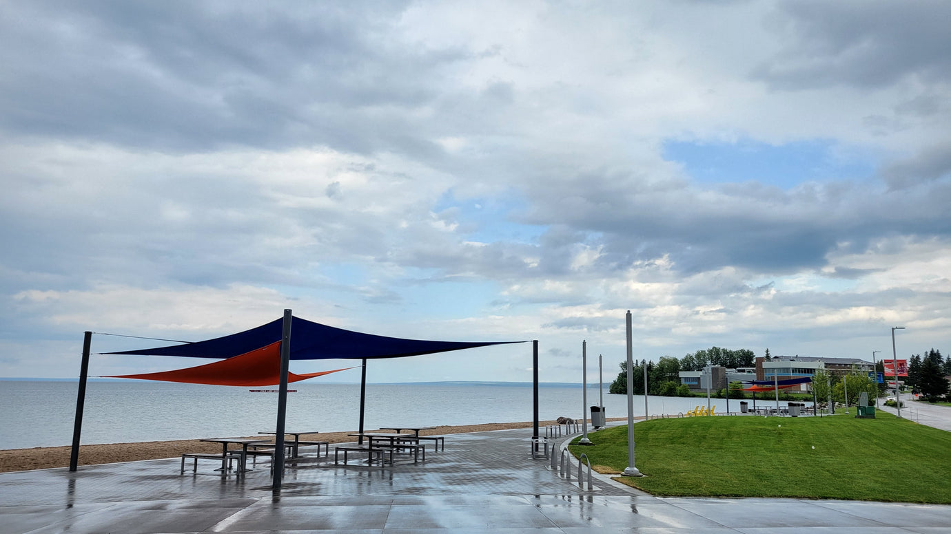 Multiple shade sails over picnic tables at a waterfront park on a rainy day, showing effective installations and precautions related to “should I take my shade sail down in a storm.”