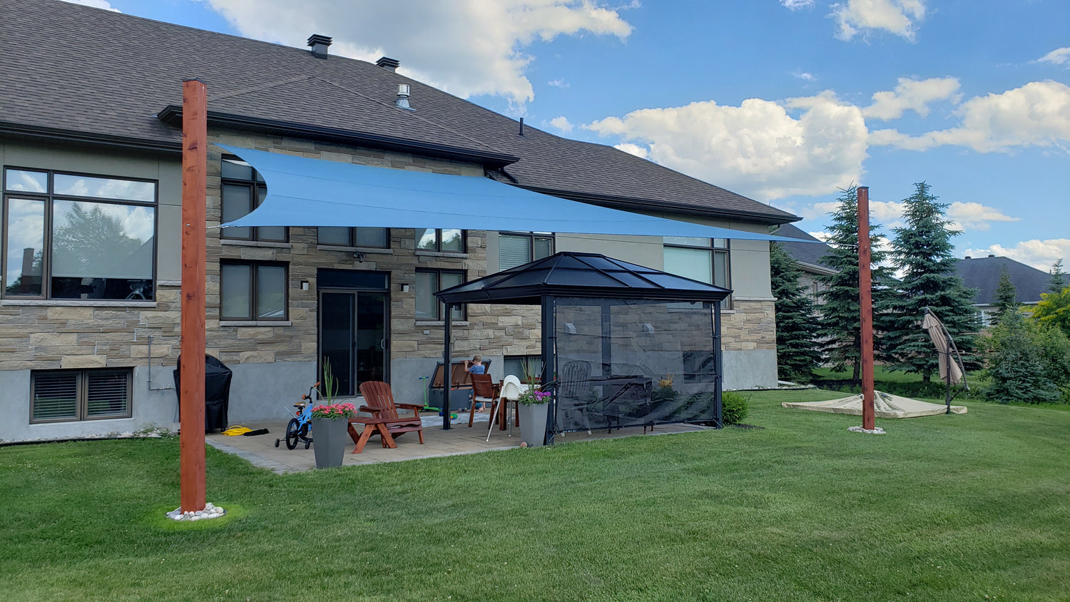 A backyard patio featuring a modern house with a blue patio shade sail stretched between two wooden posts, providing shade over a seating area with Adirondack chairs and a gazebo, illustrating the practical and aesthetic benefits of patio shade sails.