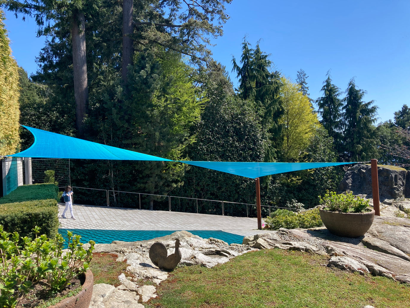  Vibrant blue canopy awnings stretch between poles, providing ample shade in a beautiful outdoor area. The awnings create a cozy space beneath, surrounded by lush greenery, tall trees, and well-maintained landscaping, enhancing the serene atmosphere.