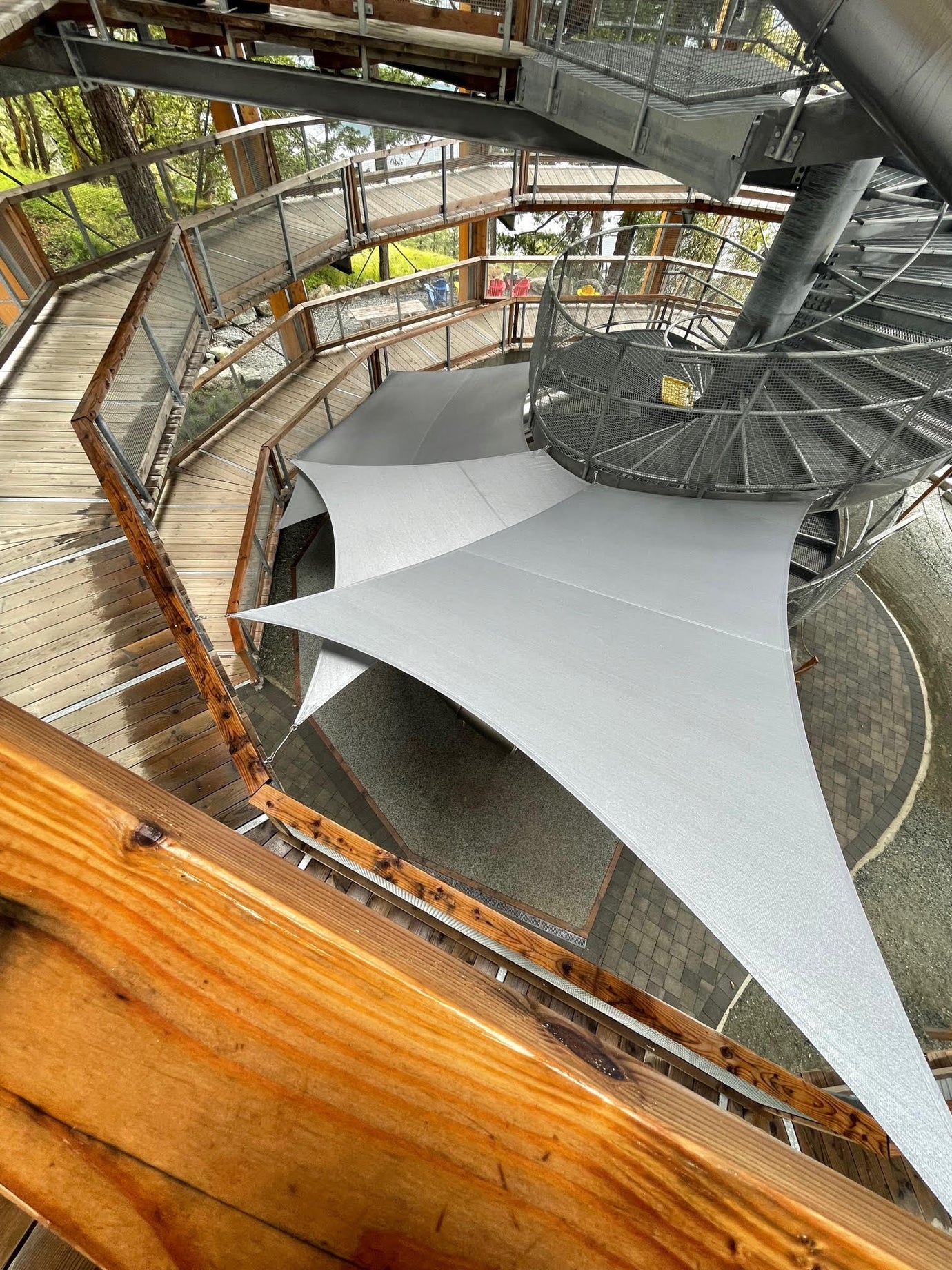 A striking spiral staircase and wooden walkways leading through a forested area, showcasing sustainable outdoor spaces with modern architectural elements and natural integration.