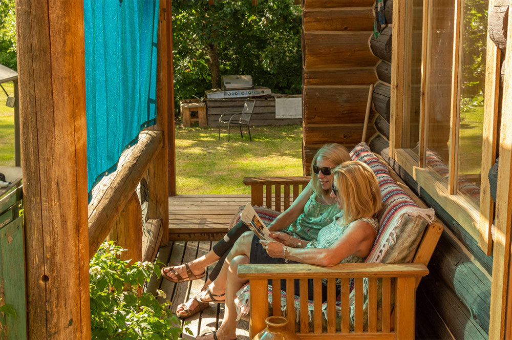 Two women relaxing on a wooden porch under a blue shade sail, enjoying the cool and comfortable atmosphere created by the shade and sails, ideal for peaceful outdoor gatherings.