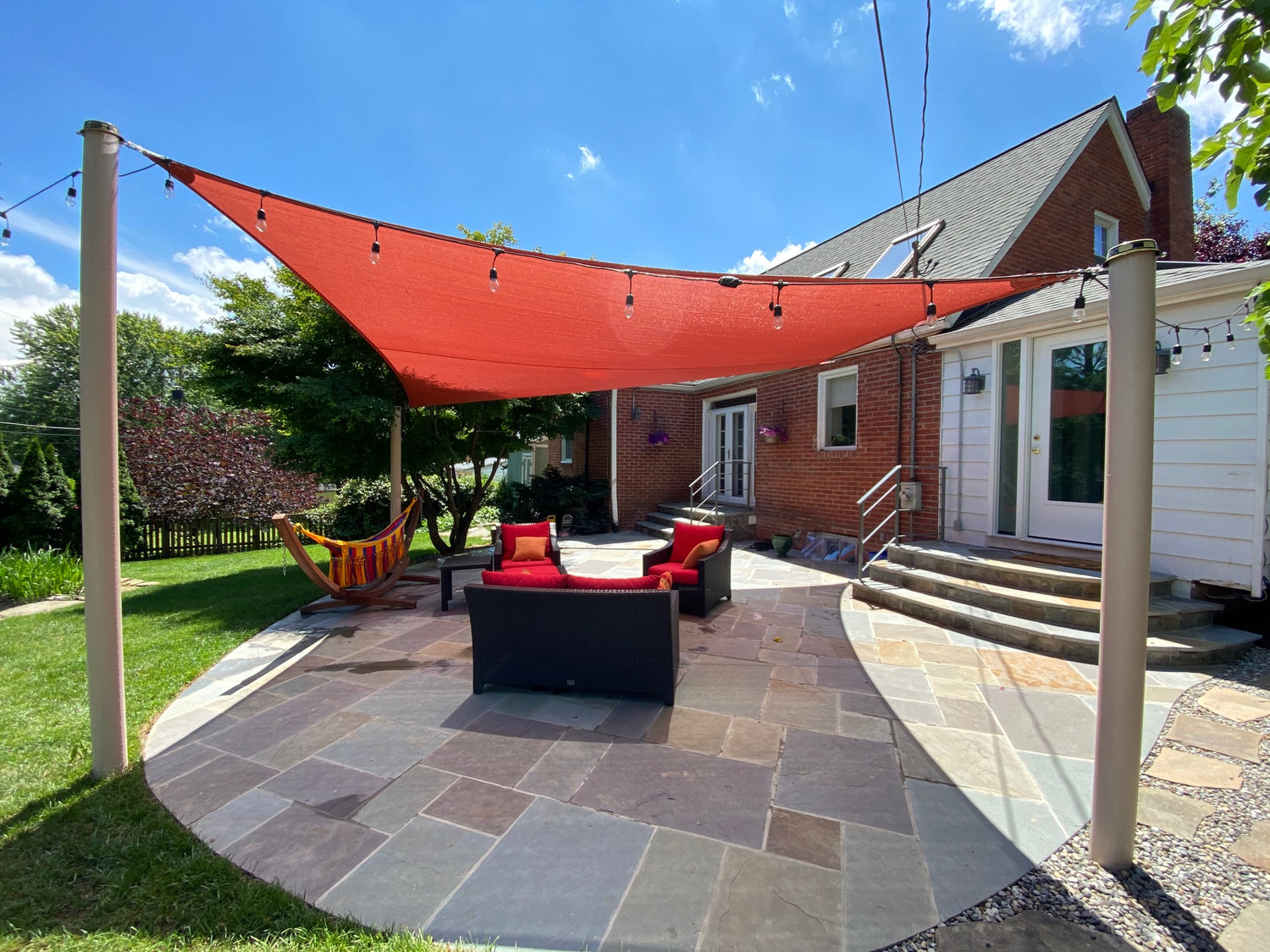 A backyard patio with a red shade sail supported by "poles for shade sails." The space features a hammock with colorful fabric, outdoor seating with red cushions, string lights, and a mix of grass and stone paving, creating a cozy and inviting atmosphere.