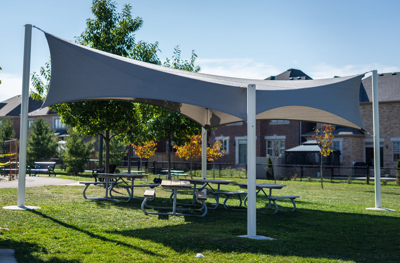Wide triangular shade cloth structure in a neighborhood park, offering shaded space above multiple picnic tables with trees and homes in the background.