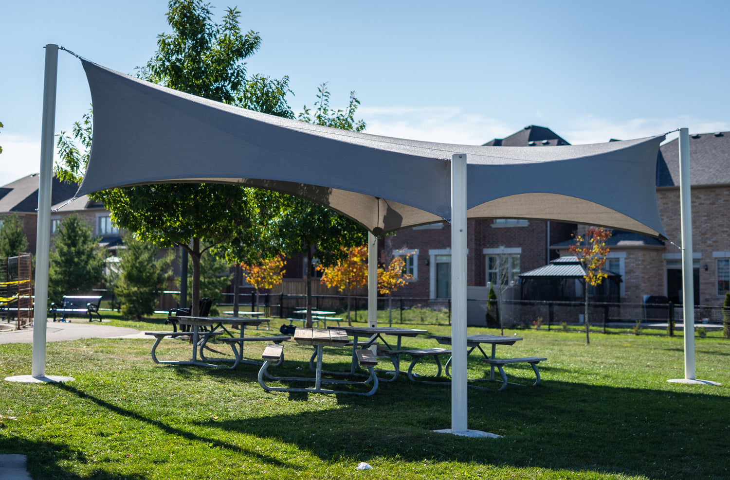 Wide triangular shade cloth structure in a neighborhood park, offering shaded space above multiple picnic tables with trees and homes in the background.
