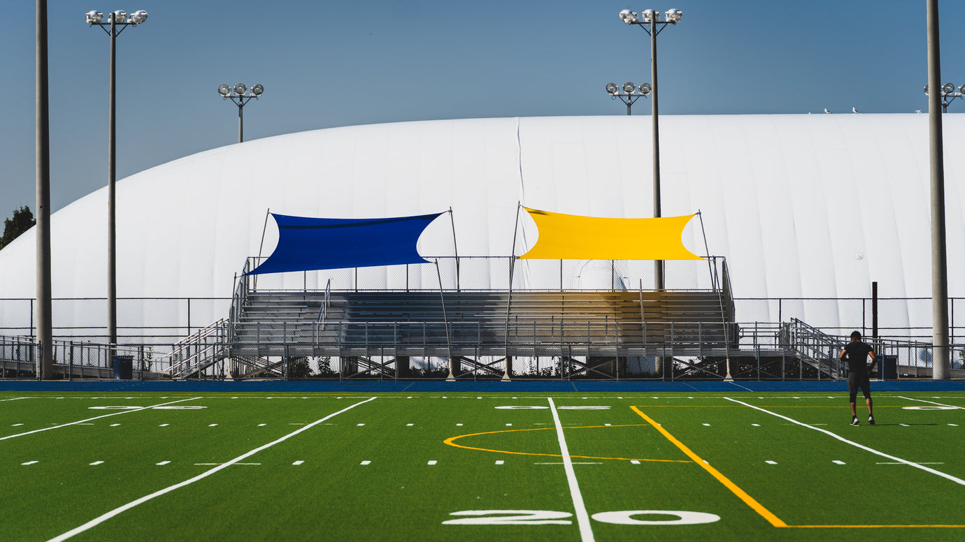 Blue and yellow Rain Shade canopy panels installed above stadium bleachers, providing weather protection and sun coverage for spectators.