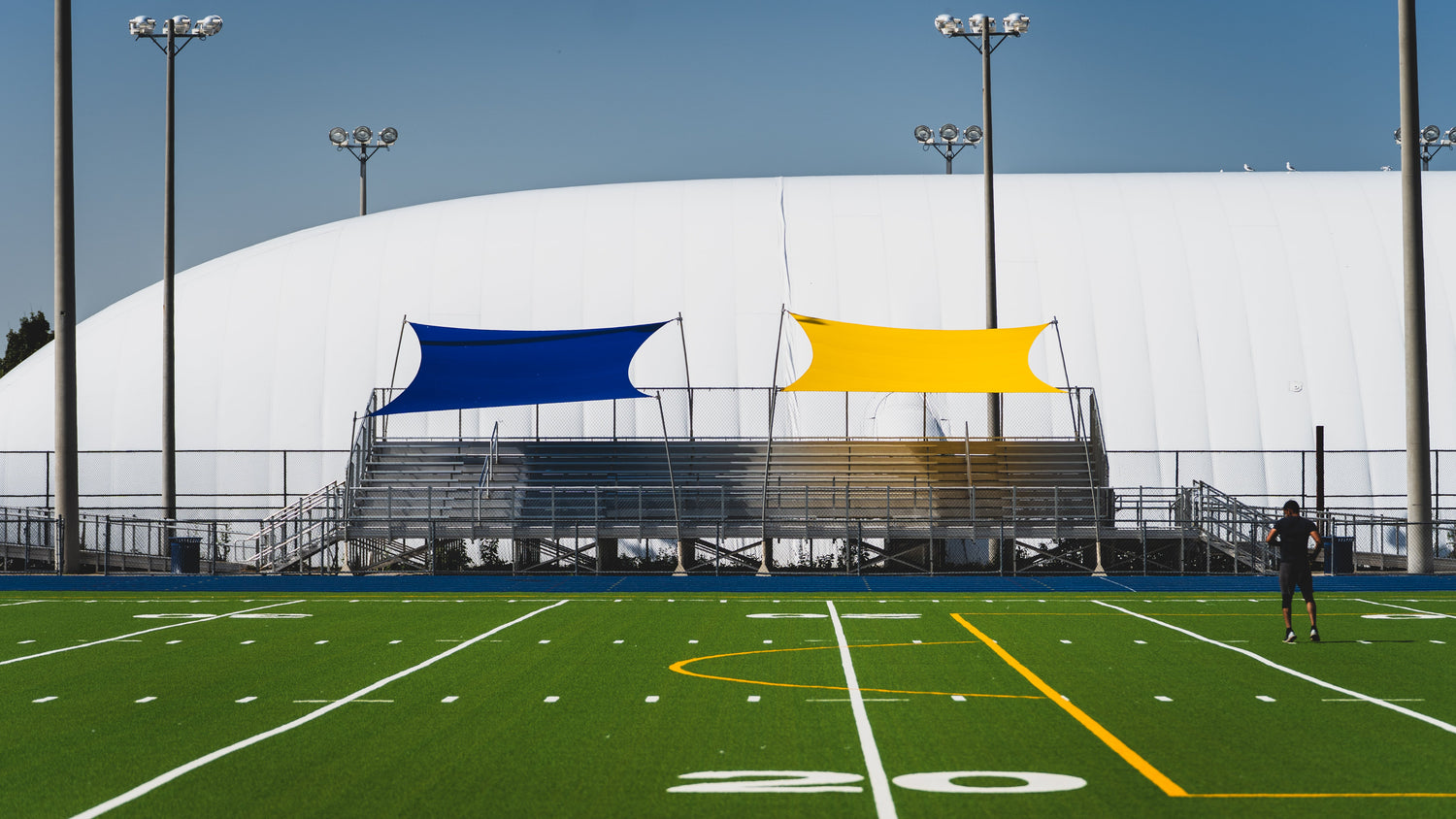 Blue and yellow Rain Shade canopy panels installed above stadium bleachers, providing weather protection and sun coverage for spectators.