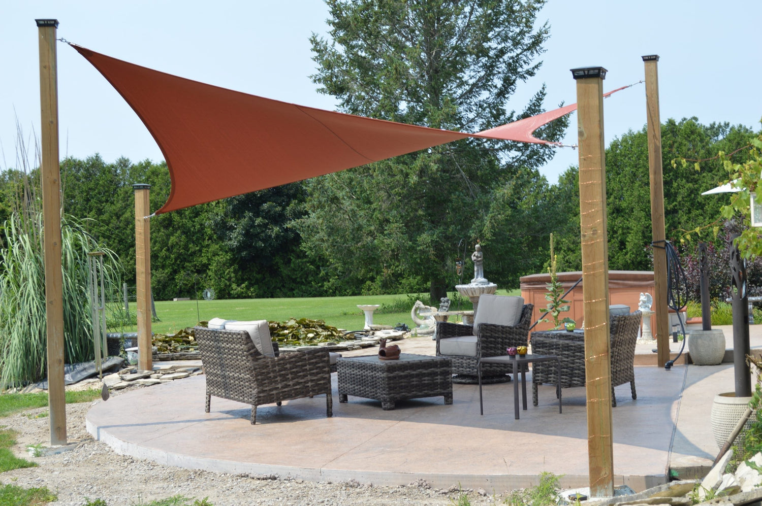 Large triangular sail stretched between posts over wicker furniture on a circular slab, showcasing a modern shade structures patio in a spacious garden.