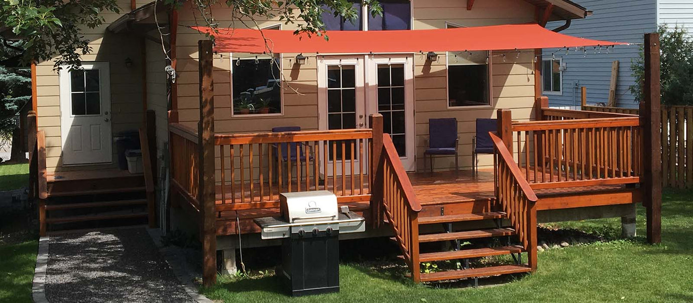 Wide-angle shot of a residential deck using a red shade sail attached to wooden posts, demonstrating how to shade a deck for backyard dining comfort.