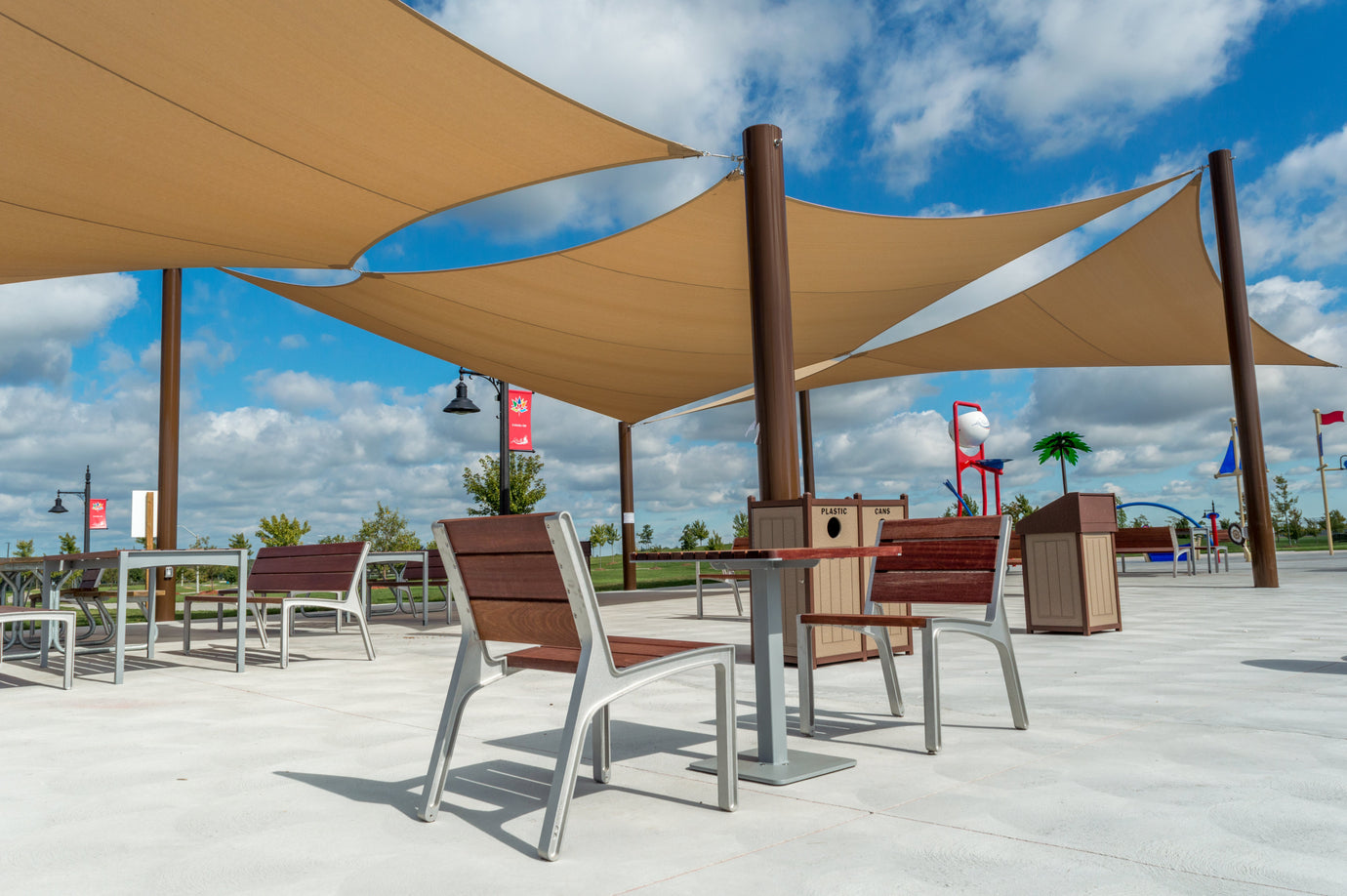 Outdoor seating area in a modern park featuring multiple beige triangular shade canopy sails mounted on tall posts, covering tables, benches, and a recycling bin under a bright blue sky with scattered clouds.