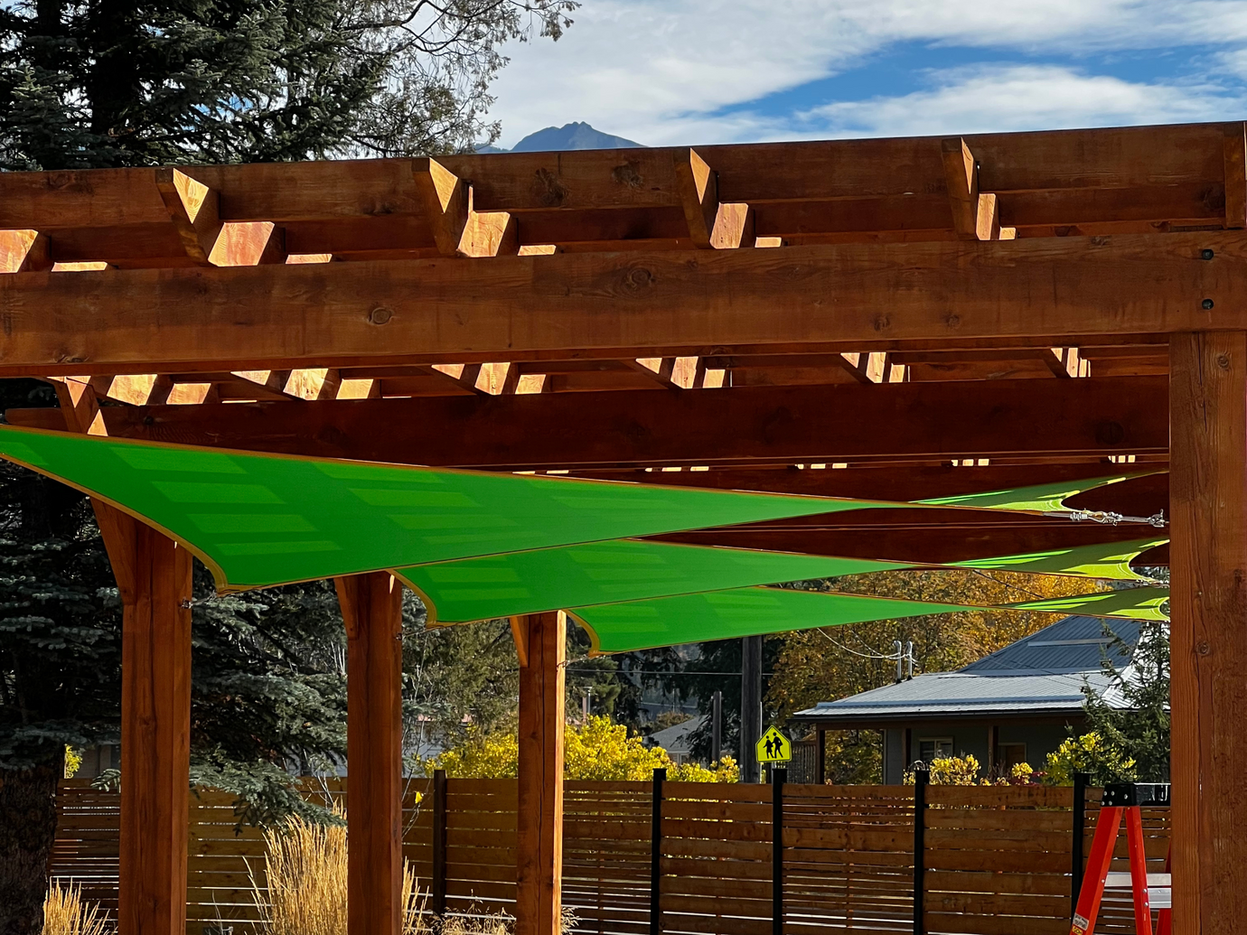  A wooden pergola with green hanging shade sails installed, creating a shaded area beneath. The background includes a clear sky and tall trees, highlighting an outdoor, nature-inspired setting.