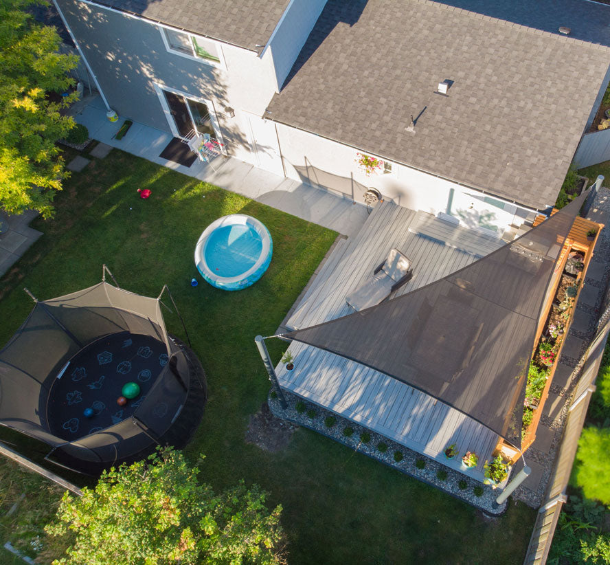 Overhead view of a backyard deck covered by a dark Shade Sail Shade, creating a cool lounging area beside a trampoline and kiddie pool.