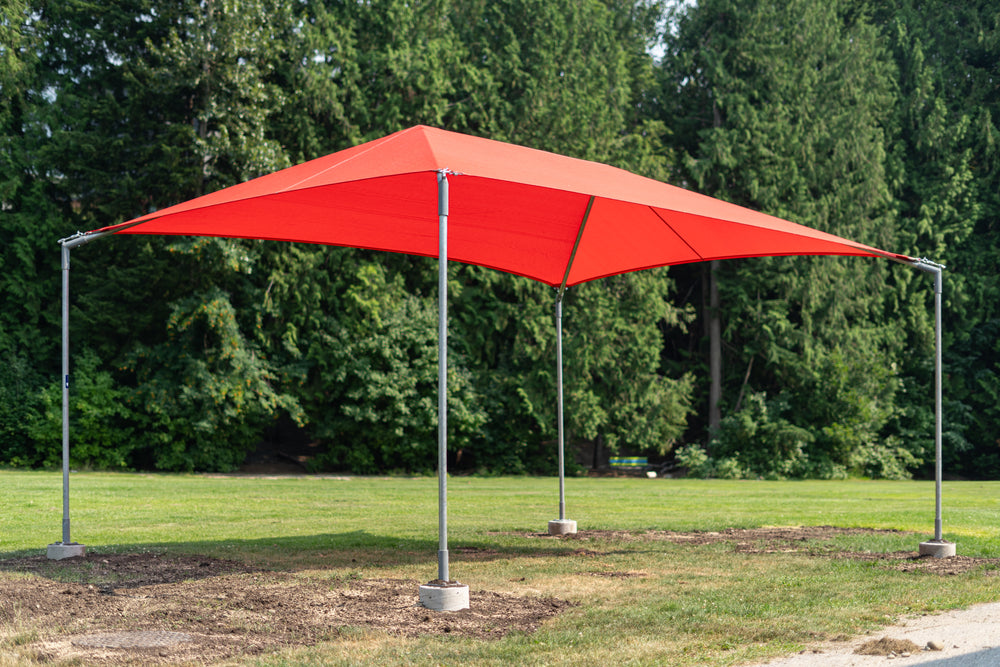 A bright red square canopy shade tent with a silver metal frame and concrete base pads, set up on a grassy lawn with a background of green trees.